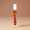A 10 milliliter clear cylindrical roller bottle filled with pale yellow perfume oil infused with dried flowers featuring a white cap and white vertical text that reads 'DEAR BOTANICS essential oil', set against a solid warm beige background.