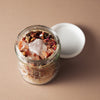 An open clear glass jar filled with a bath or body scrub, topped with dried flowers and crystals, with its white lid resting beside it on a warm beige background.