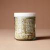 A clear 220 gram glass jar with the ingredients in white lettering filled with coarse botanical salt or scrub, sealed with a white lid, standing on a warm beige background. 