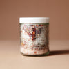 A 220 gram glass jar with white letters of ingredients with a white lid containing a mix of dried flowers, salts and crystals on a beige background.