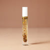 A 10 milliliter clear cylindrical roller bottle filled with pale yellow perfume oil infused with dried flowers featuring a white cap and white vertical text that reads 'DEAR BOTANICS essential oil', set against a solid warm beige background.