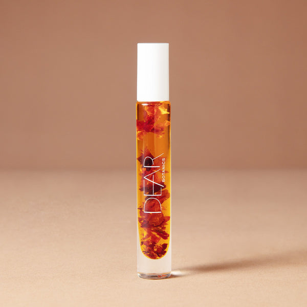 A 10 milliliter clear cylindrical roller bottle filled with pale yellow perfume oil infused with dried flowers featuring a white cap and white vertical text that reads 'DEAR BOTANICS essential oil', set against a solid warm beige background.