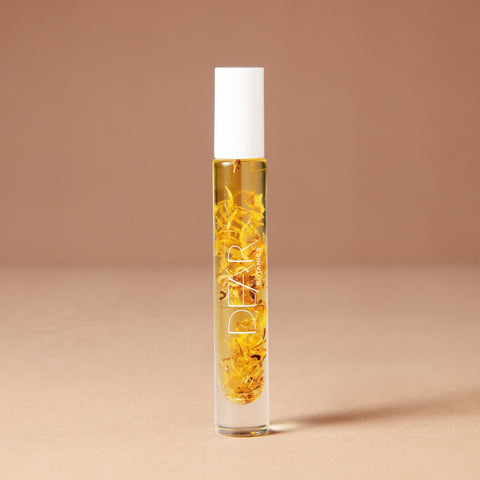 A 10 milliliter clear cylindrical roller bottle filled with pale yellow perfume oil infused with dried flowers featuring a white cap and white vertical text that reads 'DEAR BOTANICS essential oil', set against a solid warm beige background.